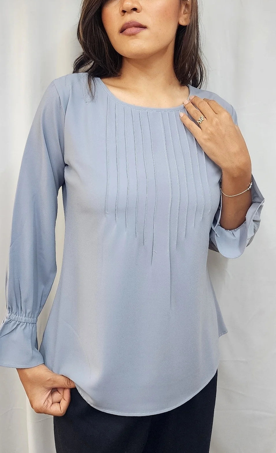 A woman modeling a light blue long-sleeve blouse with vertical pleats on the bodice and gathered sleeves, paired with dark pants, against a light gray fabric backdrop.