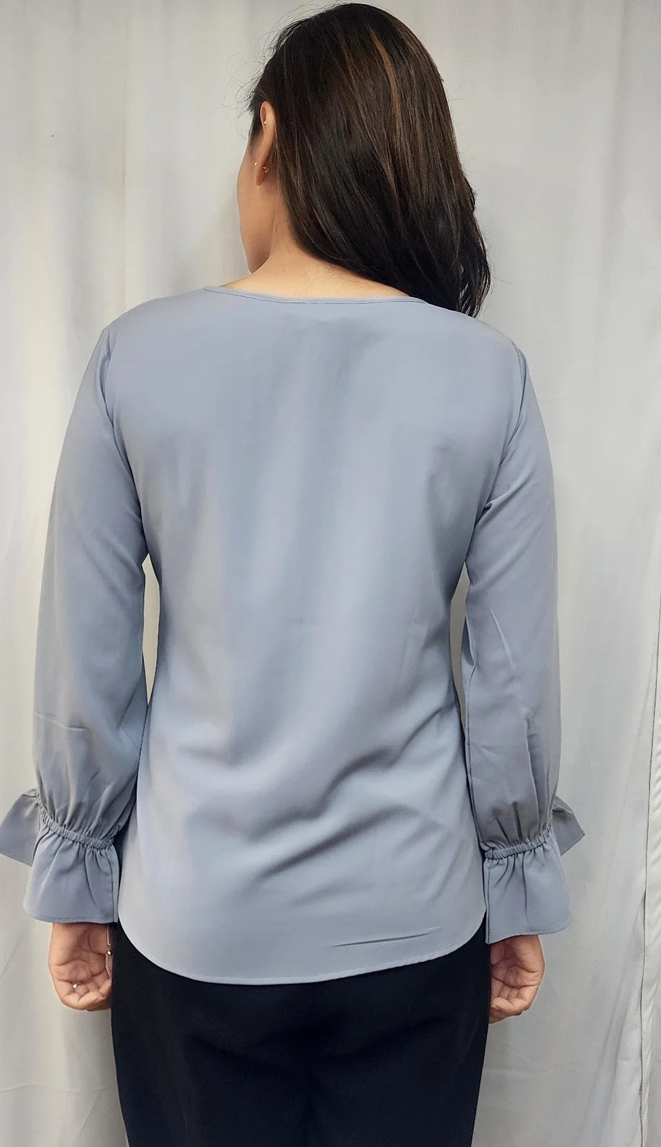 A woman viewed from the back wearing a light blue-gray long-sleeve blouse with ruffled cuffs and dark pants, standing in front of a white curtain.