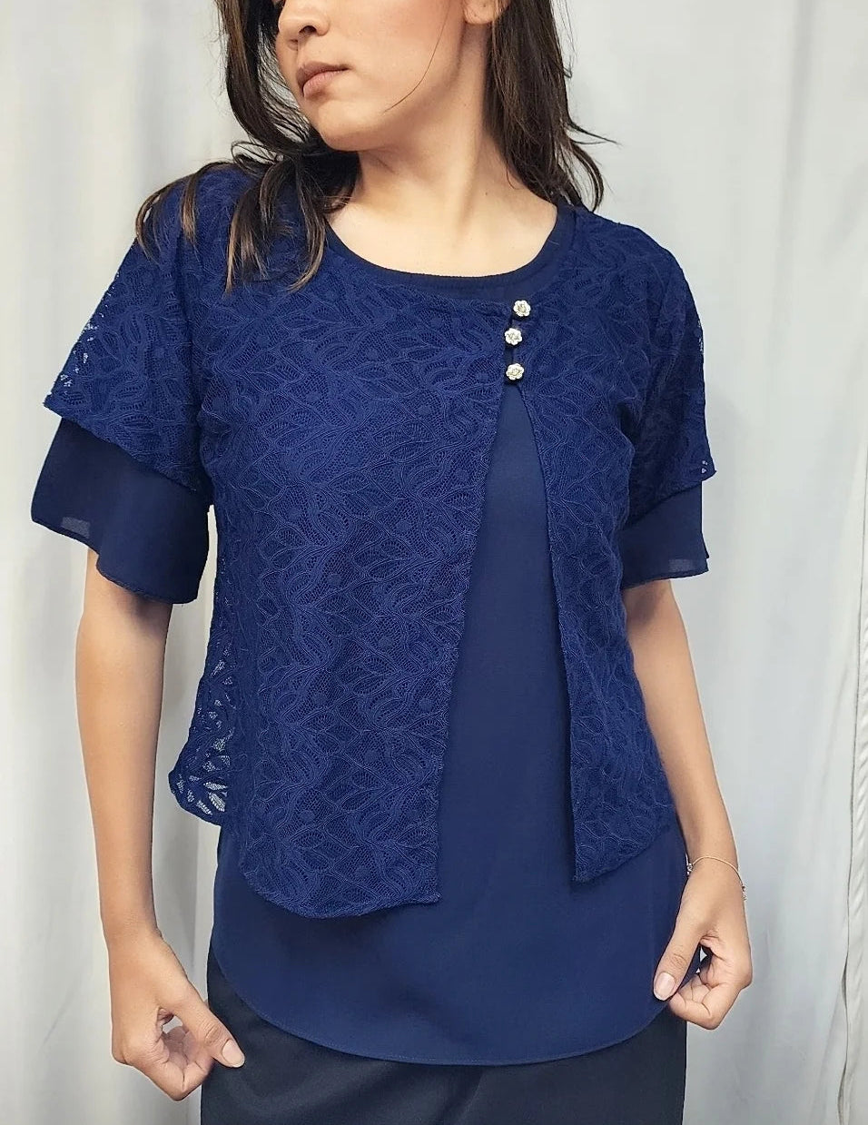 A woman wearing a navy blue short-sleeve blouse with a textured lace overlay and three decorative buttons on the chest, paired with a matching navy top underneath.