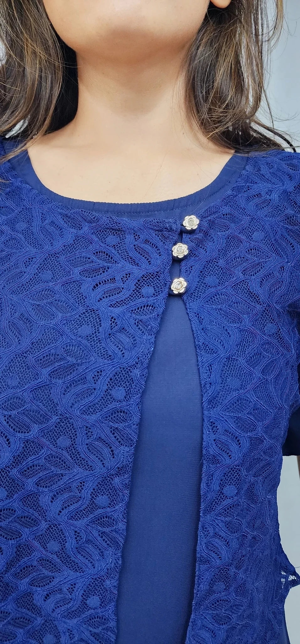 Close-up of a person wearing a royal blue lace top with a solid blue vertical panel and three decorative silver-tone embellishments at the neckline.