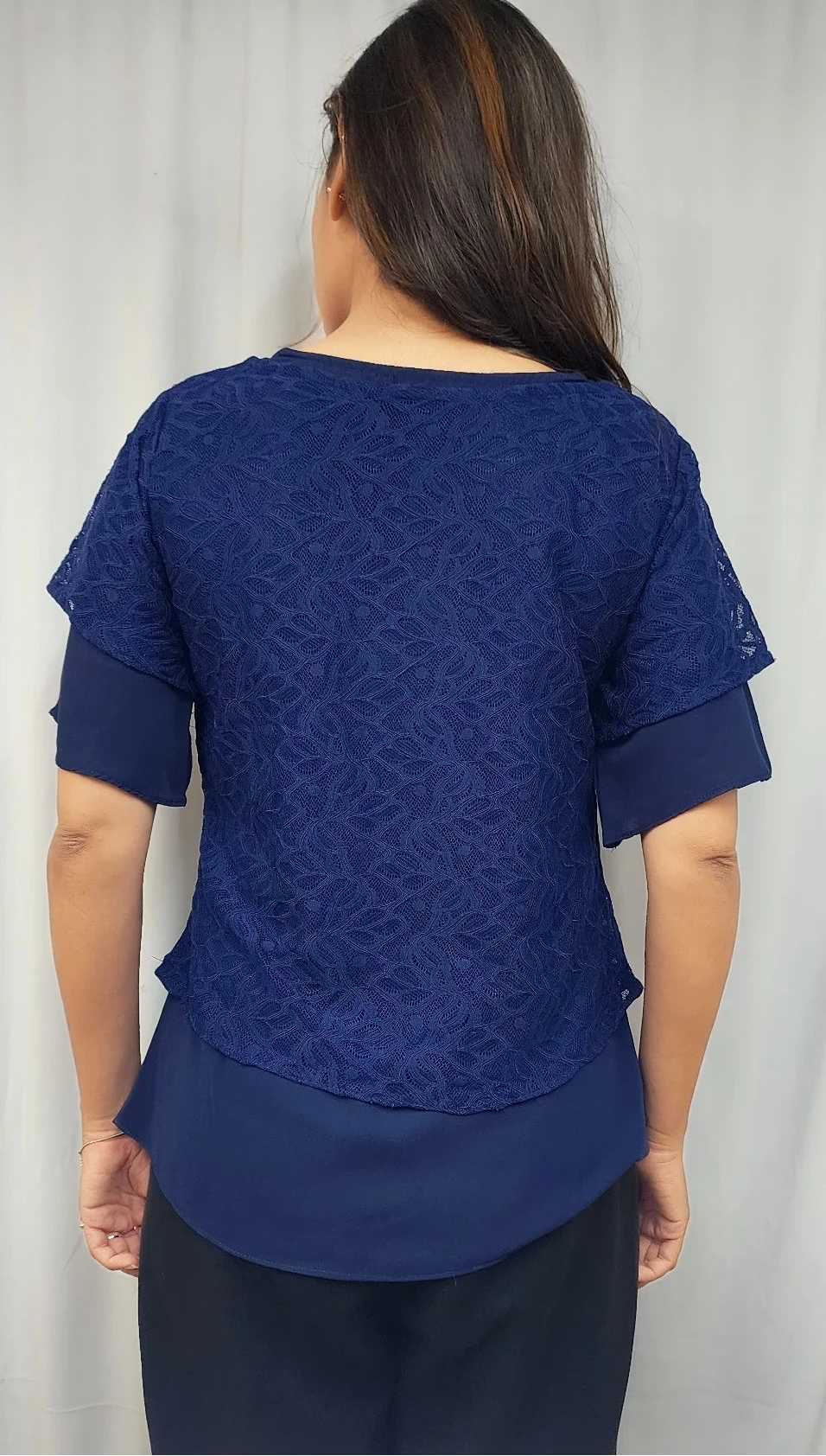 A woman viewed from the back wearing a royal blue short-sleeve top with a textured lace back panel and solid blue layered sleeves, paired with dark navy pants against a light gray curtain background.