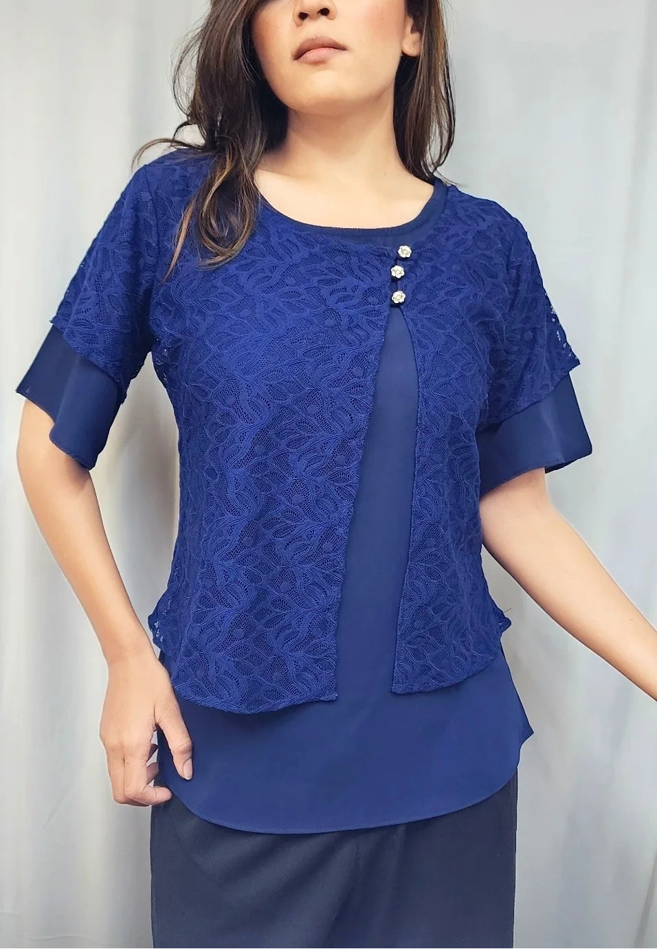 A woman modeling a royal blue lace short-sleeve blouse with three decorative buttons on the chest, paired with dark navy pants, set against a light gray curtain backdrop.