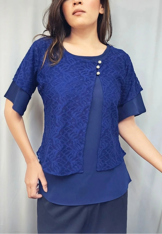A woman modeling a royal blue lace short-sleeve blouse with three decorative buttons on the chest, paired with dark navy pants, set against a light gray curtain backdrop.
