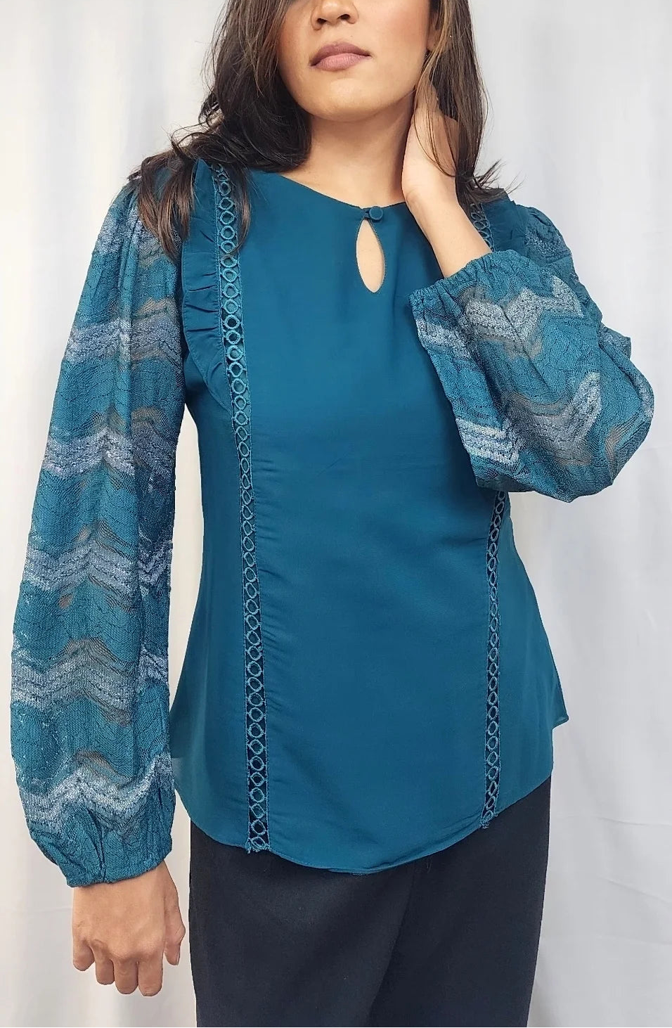 A woman modeling a teal blue blouse with patterned sheer sleeves and a keyhole neckline, paired with dark pants, standing against a white curtain backdrop.