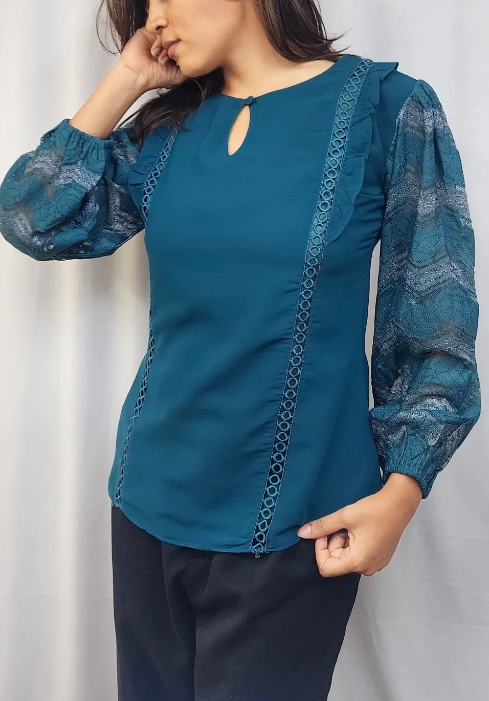 A woman wearing a teal blue long-sleeve blouse with patterned sheer sleeves and a keyhole neckline, paired with dark pants, standing in front of a white curtain.