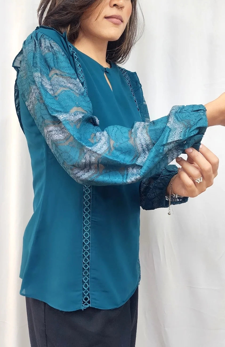 A woman wearing a teal blue long-sleeve blouse with sheer patterned sleeves and dark pants, standing against a white curtain backdrop.