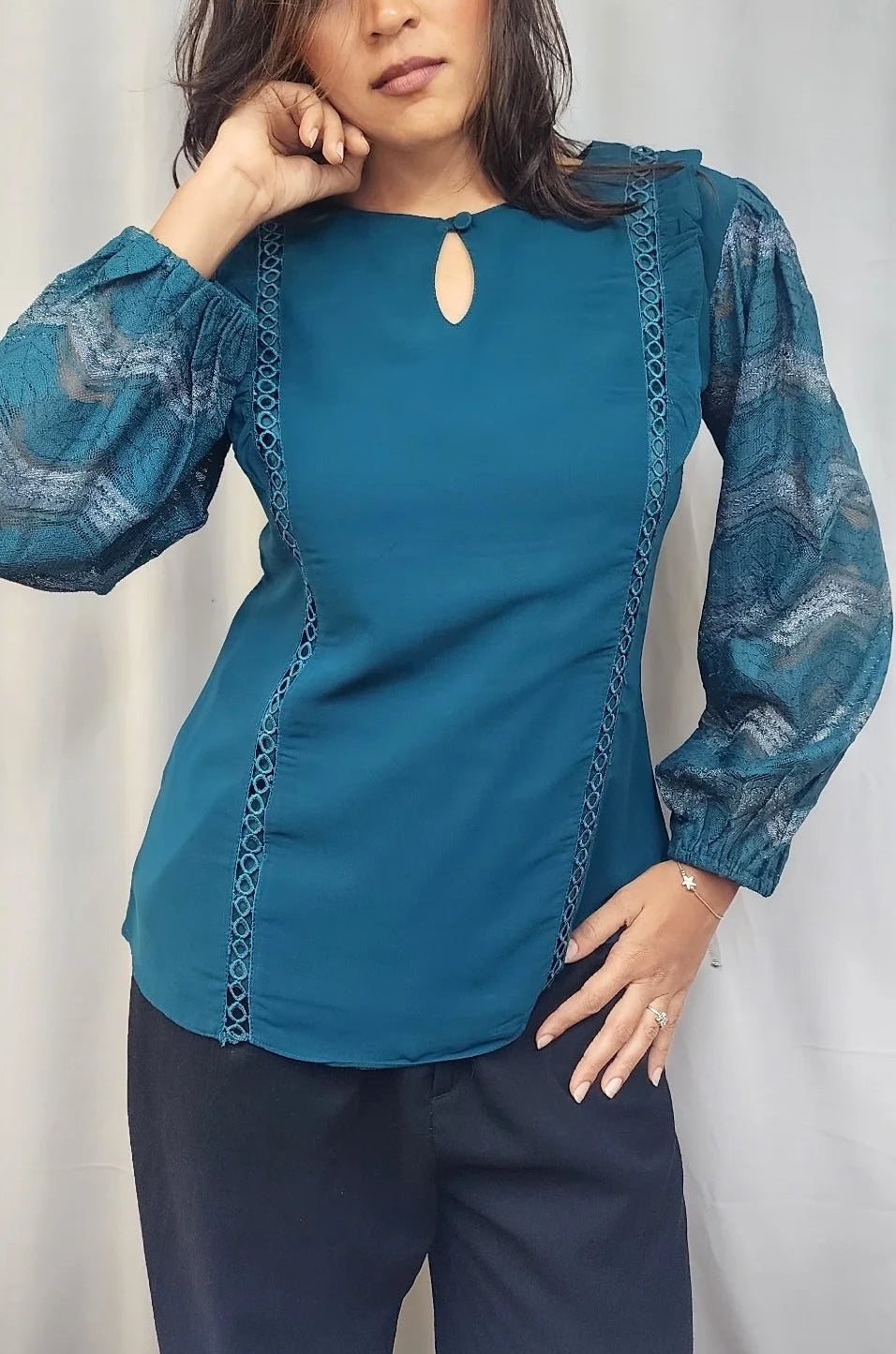 A woman modeling a teal blue long-sleeve blouse with sheer patterned sleeves and a keyhole neckline, paired with dark navy pants, against a light gray curtain backdrop.