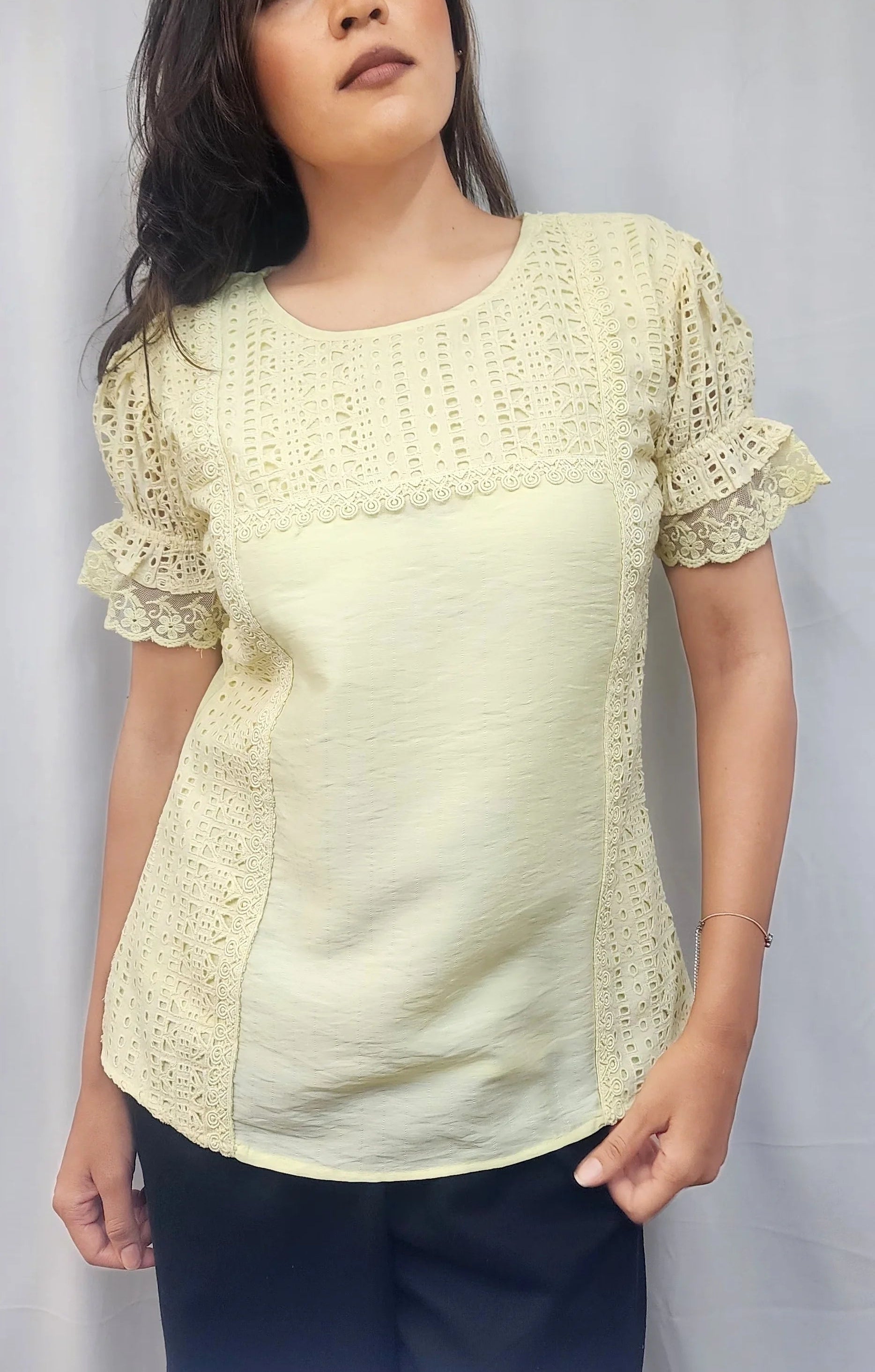 A woman wearing a pale yellow short-sleeve blouse with lace puff sleeves and a textured embroidered bodice, paired with dark pants, standing against a light gray backdrop.