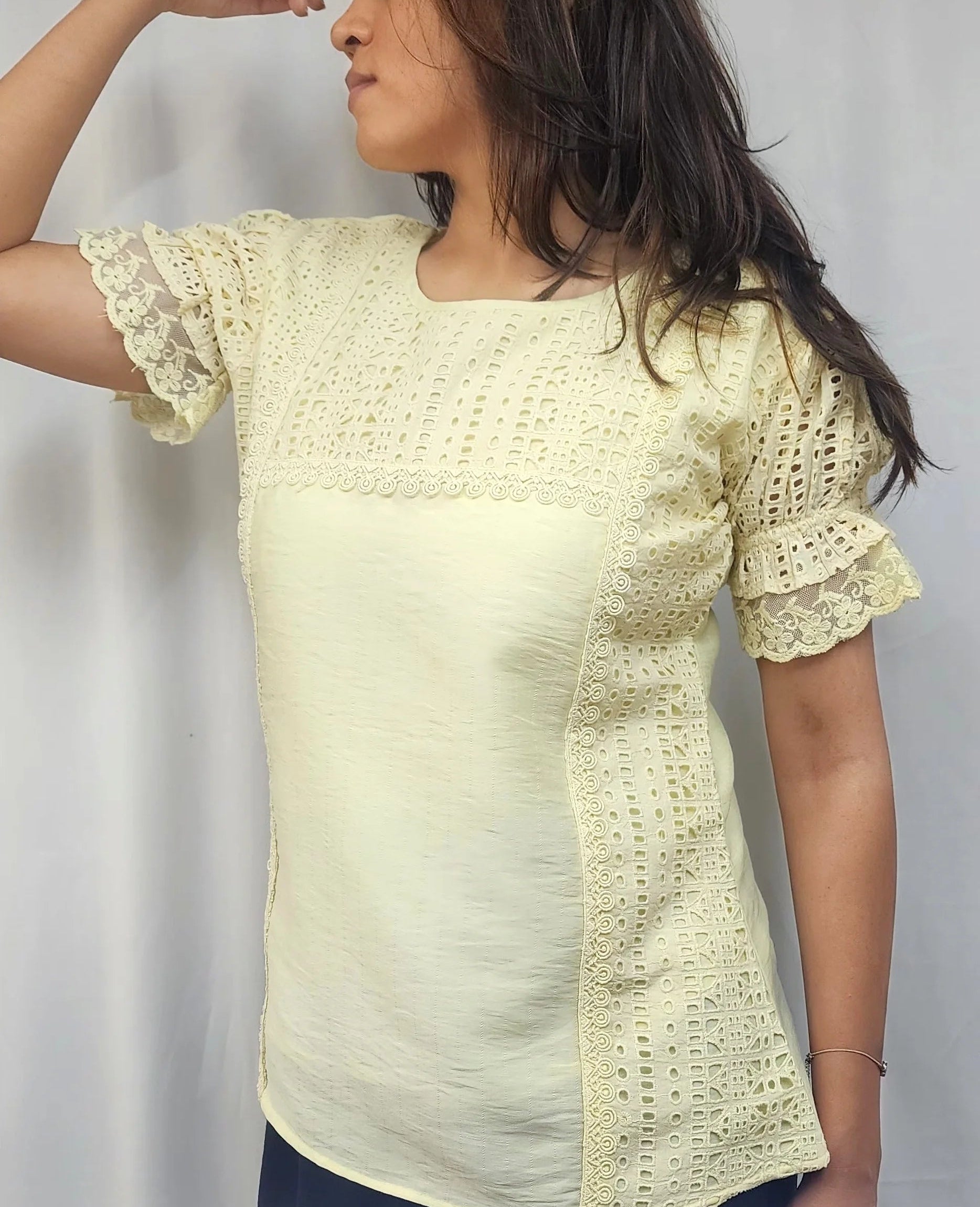 A woman wearing a pale yellow short-sleeve blouse with textured crochet lace panels on the chest and sides, paired with dark pants, standing against a white curtain background.