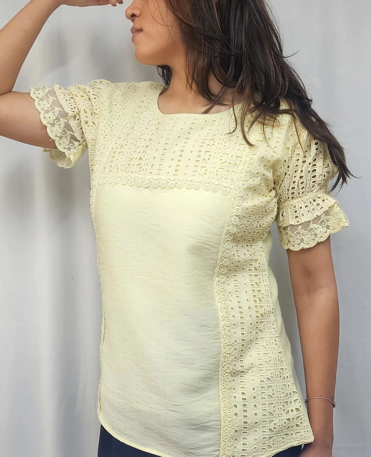 A woman wearing a pale yellow short-sleeve blouse with textured crochet lace panels on the chest and sides, paired with dark pants, standing against a white curtain background.