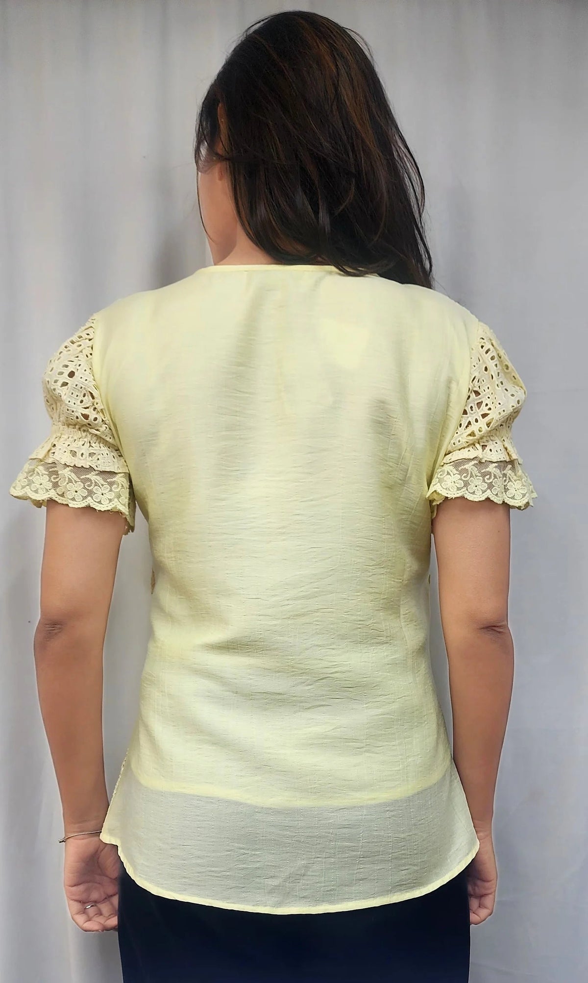 A woman viewed from behind wearing a pale yellow short-sleeve blouse with lace ruffle sleeves and a sheer underlayer, paired with dark navy bottoms, against a light gray curtain background.