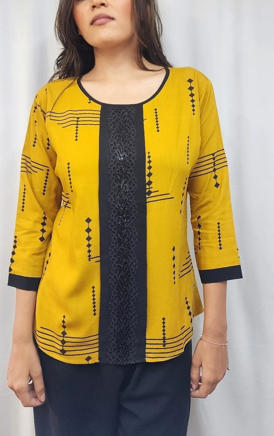 A woman modeling a mustard yellow three-quarter sleeve kurta with black geometric patterns and a black textured center panel, paired with dark navy trousers, set against a white curtain backdrop.