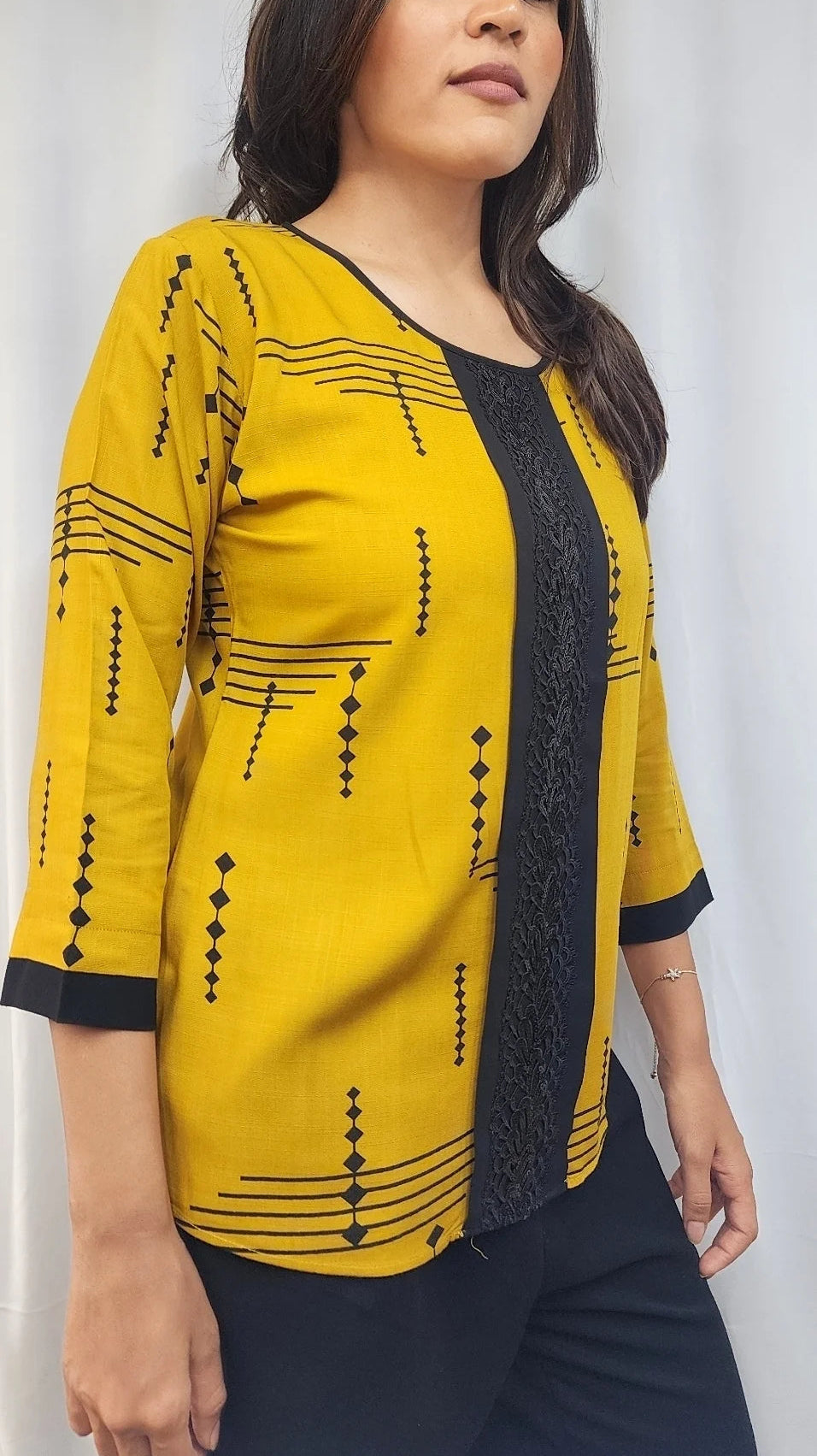 A woman wearing a mustard yellow three-quarter sleeve top with black geometric patterns and a black lace front panel, paired with dark pants, standing against a white curtain background.