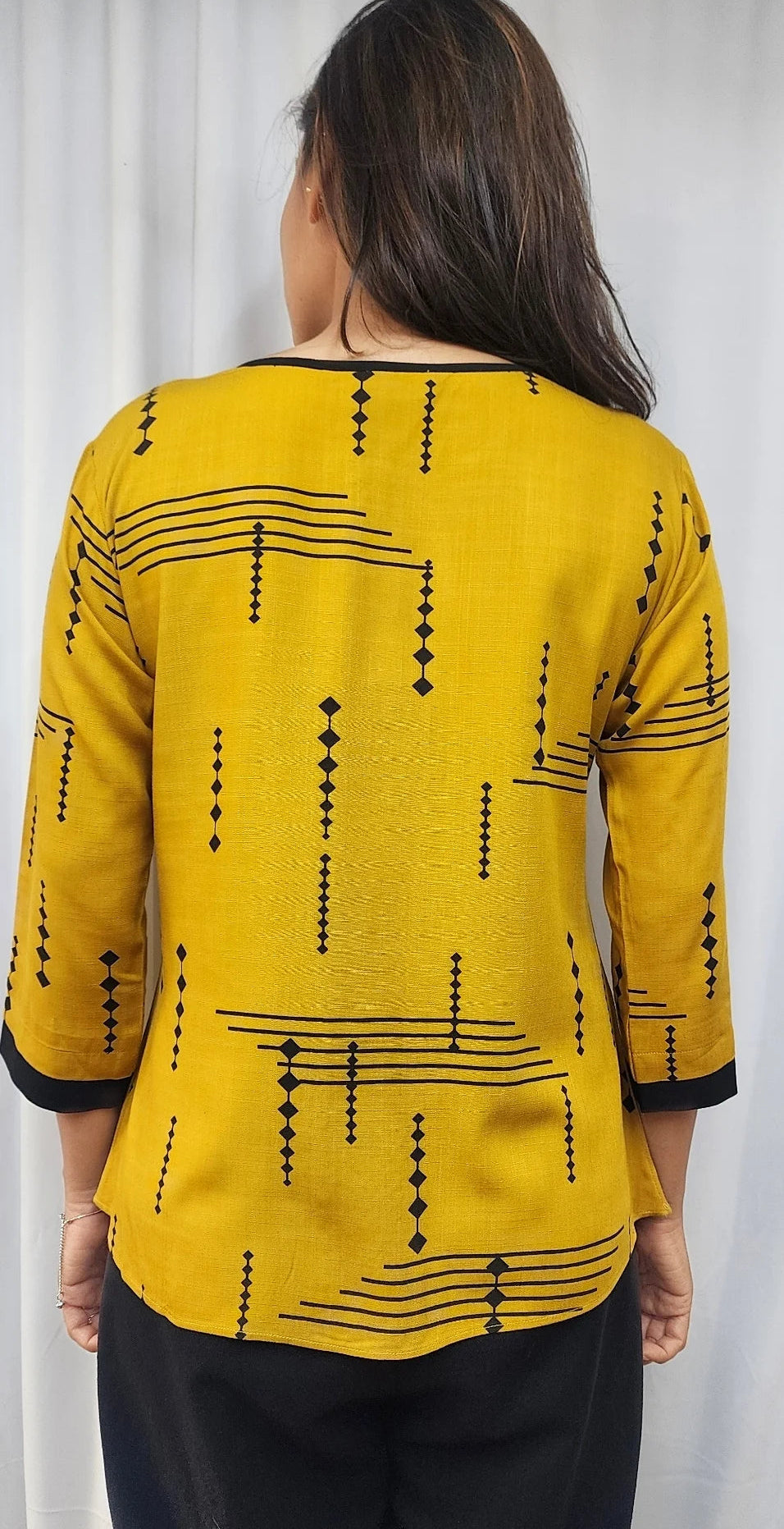 A woman viewed from the back wearing a bright yellow top with black geometric patterns and black pants, standing against a light curtain background.