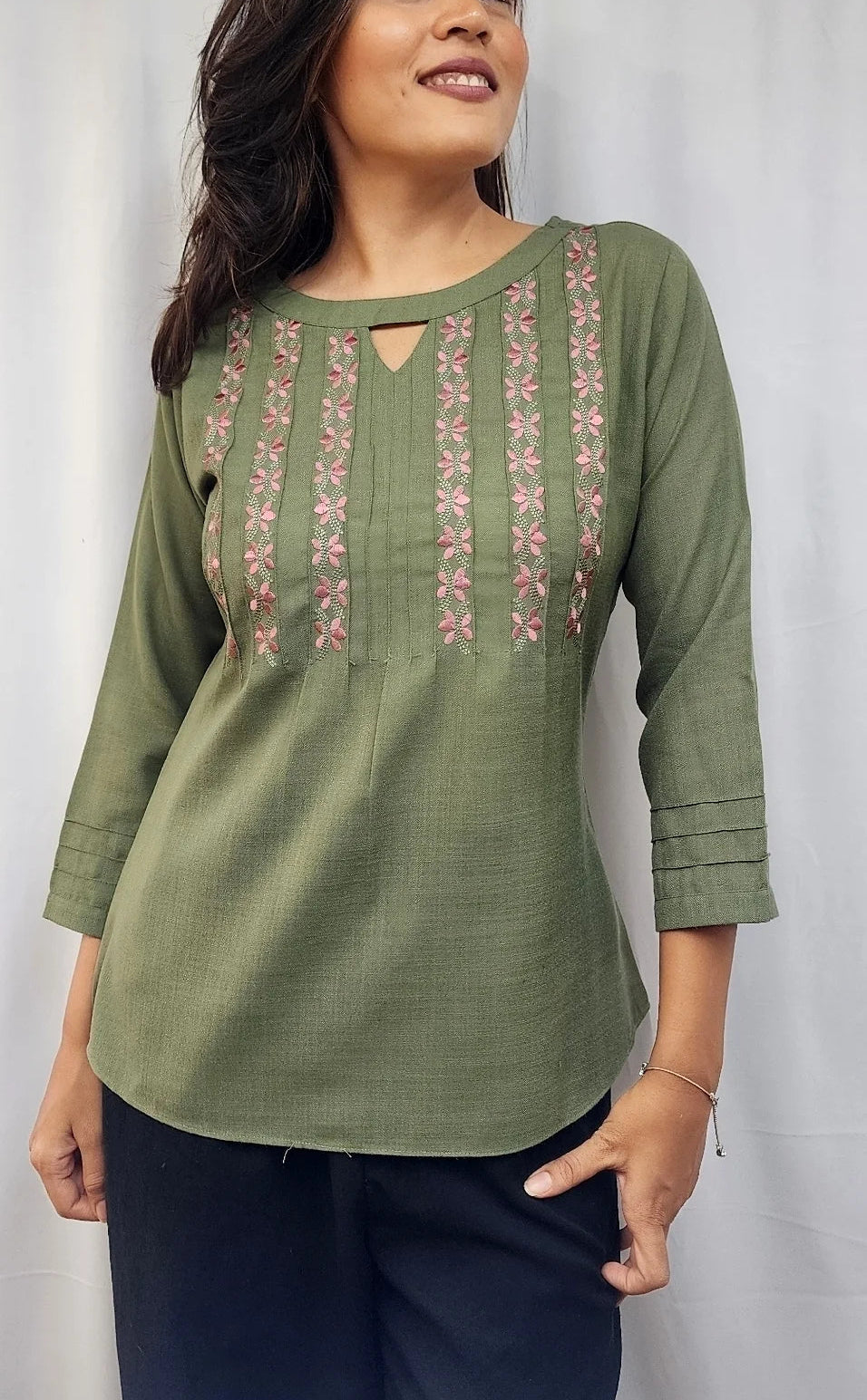 A woman wearing an olive green three-quarter sleeve kurta with vertical pink and gold embroidered patterns on the chest, paired with dark navy pants, standing against a light gray backdrop.