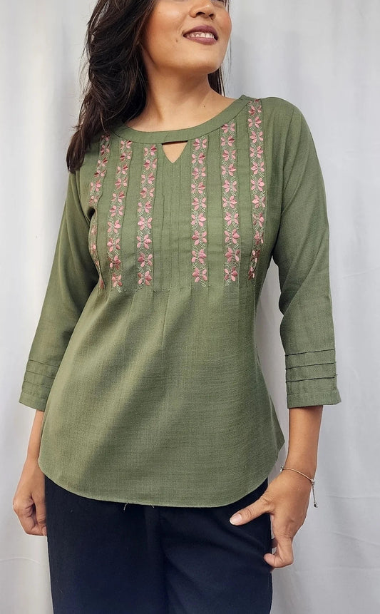A woman wearing an olive green three-quarter sleeve kurta with vertical pink and gold embroidered patterns on the chest, paired with dark navy pants, standing against a light gray backdrop.
