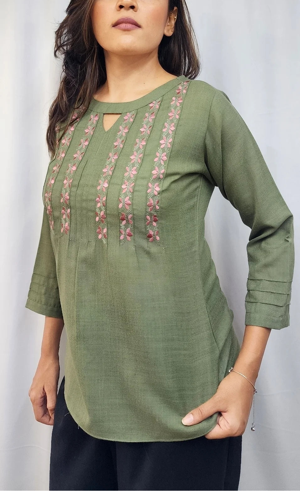 A woman modeling an olive green three-quarter sleeve kurta with vertical floral embroidery on the chest, paired with dark pants, set against a white curtain background.