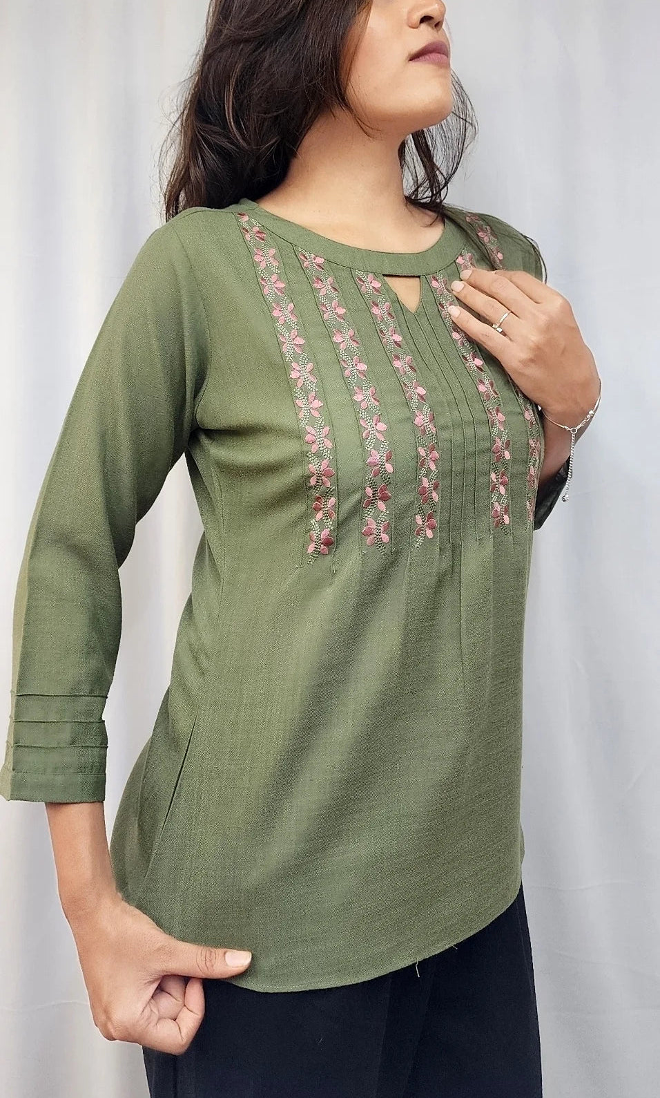 A woman wearing an olive green three-quarter sleeve kurta with vertical pink floral embroidery on the chest, paired with dark navy pants, standing in front of a white curtain.