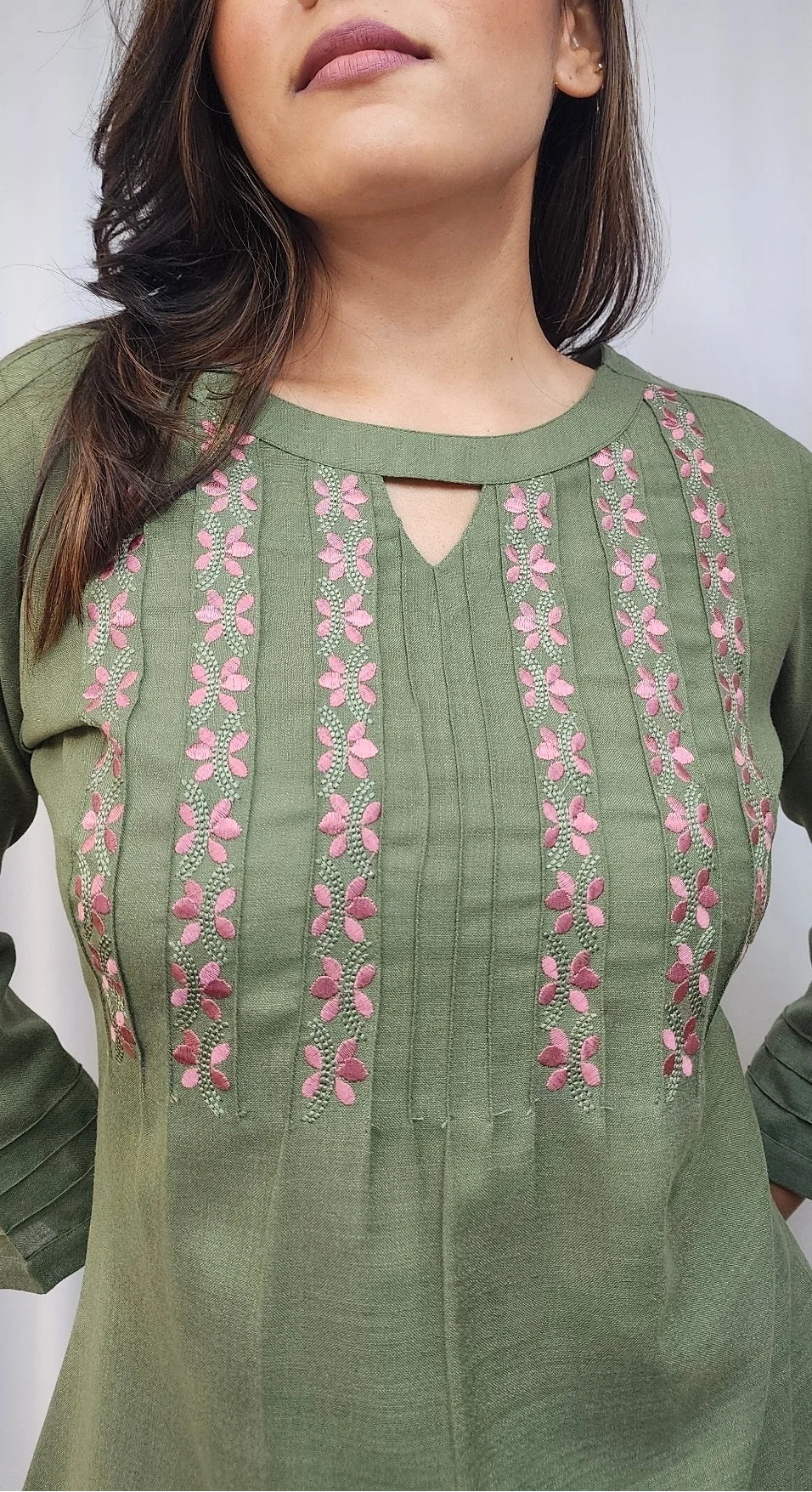 A woman wearing an olive green long-sleeve kurta with pink and red floral embroidery on the chest, photographed against a white curtain background.