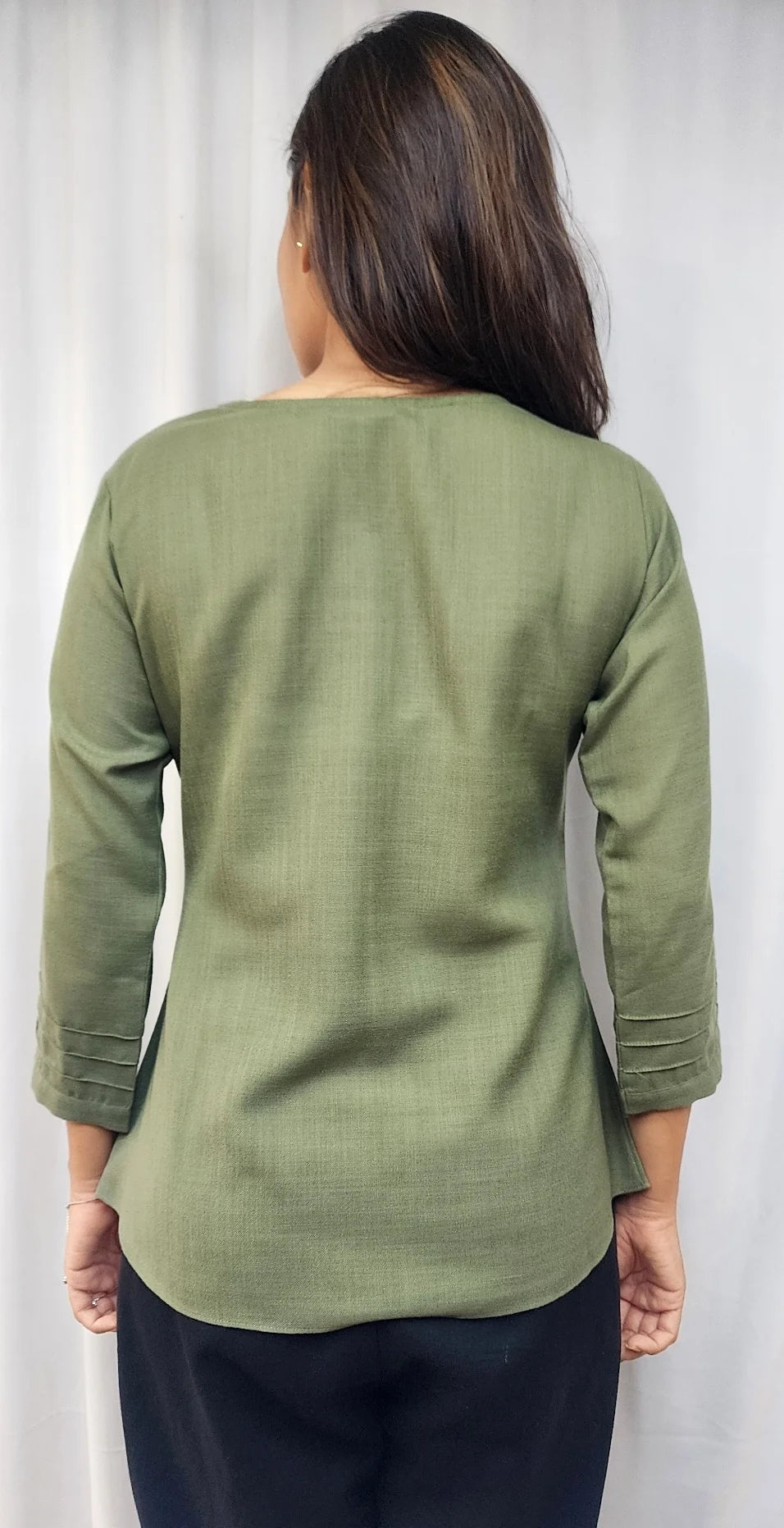 A woman viewed from the back wearing a sage green three-quarter sleeve top and dark navy pants, standing against a light gray curtain backdrop.