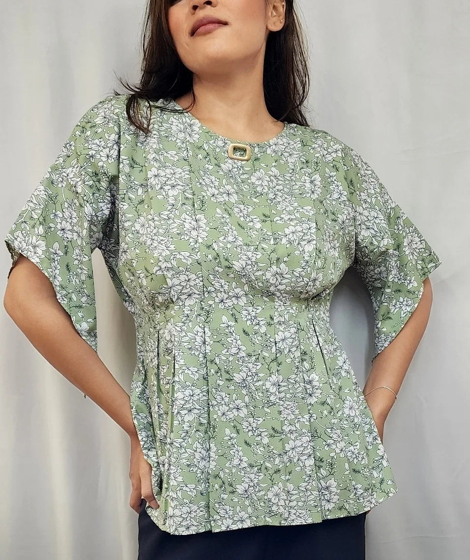 A woman wearing a light green and white floral print peplum blouse with short flutter sleeves, standing against a light gray curtain background.