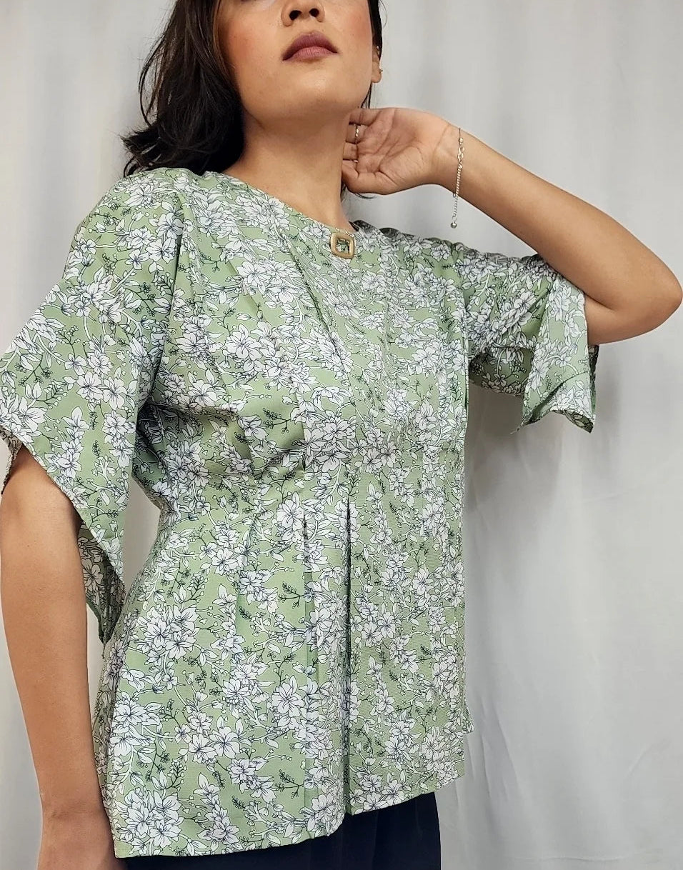 A woman wearing a light green short-sleeve blouse with a white floral pattern, standing against a white curtain backdrop.