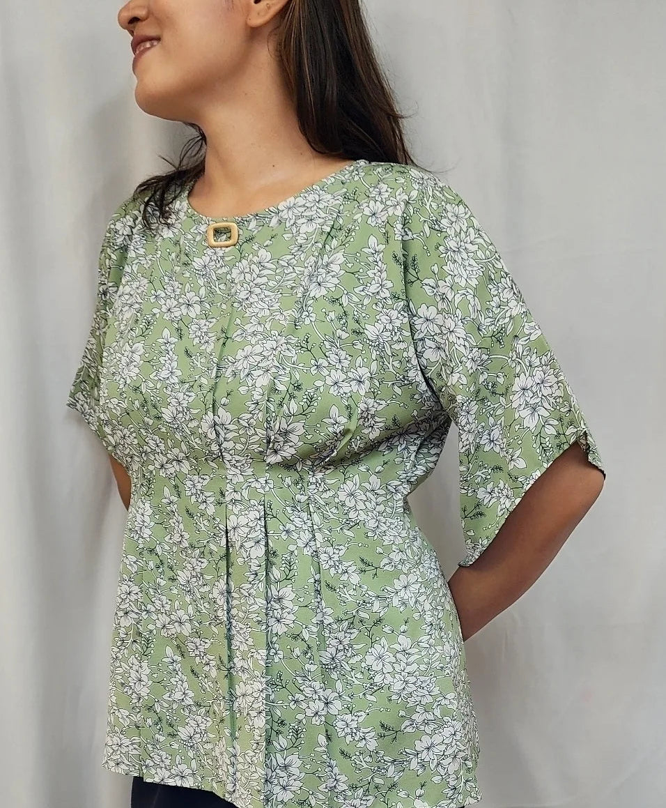 A woman wearing a light green and white floral print short-sleeve top with a gold button closure at the neckline, standing against a light gray curtain backdrop.