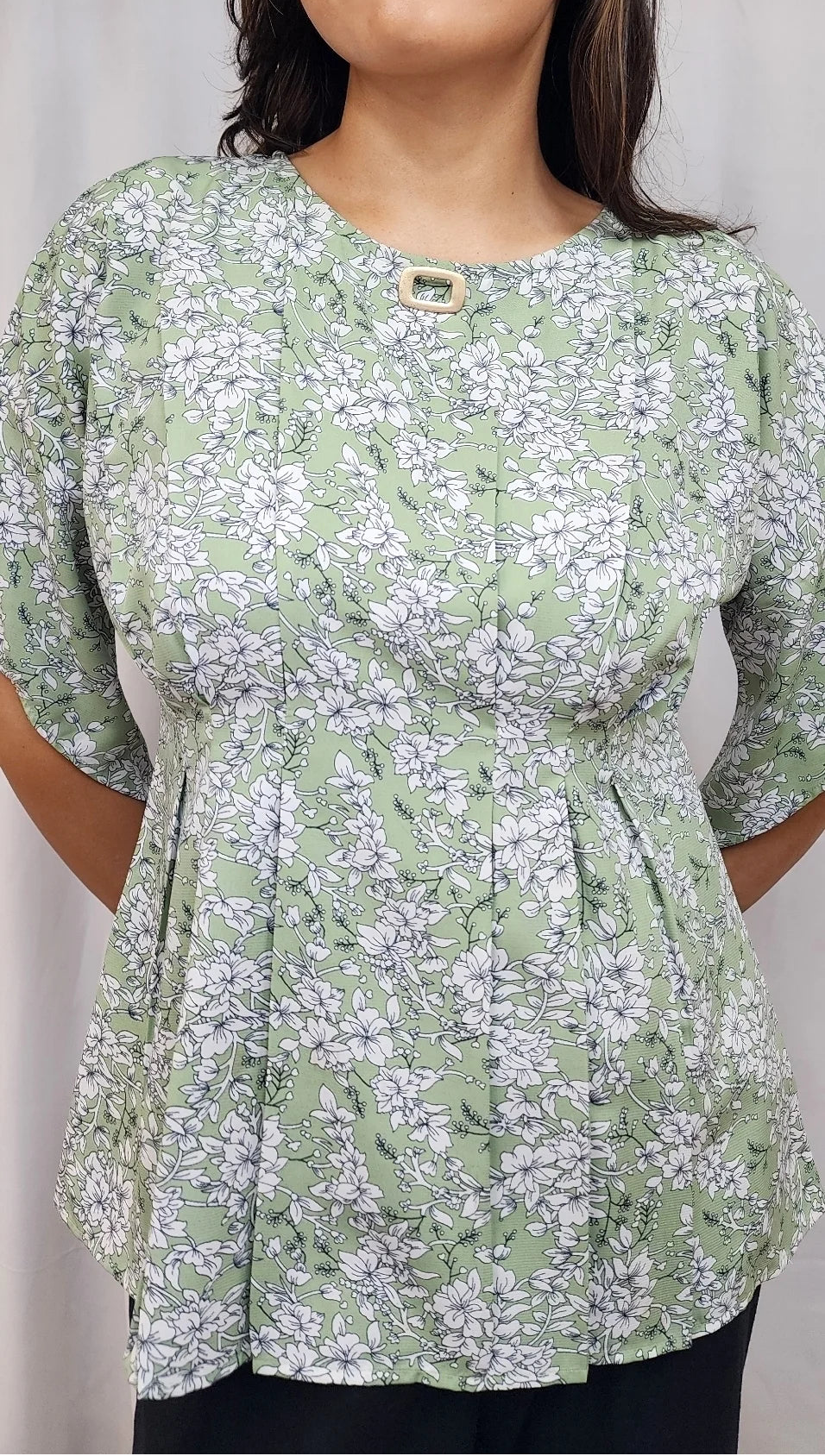 A woman wearing a light green and white floral print peplum top with three-quarter sleeves and a small gold button at the neckline, set against a neutral light gray background.