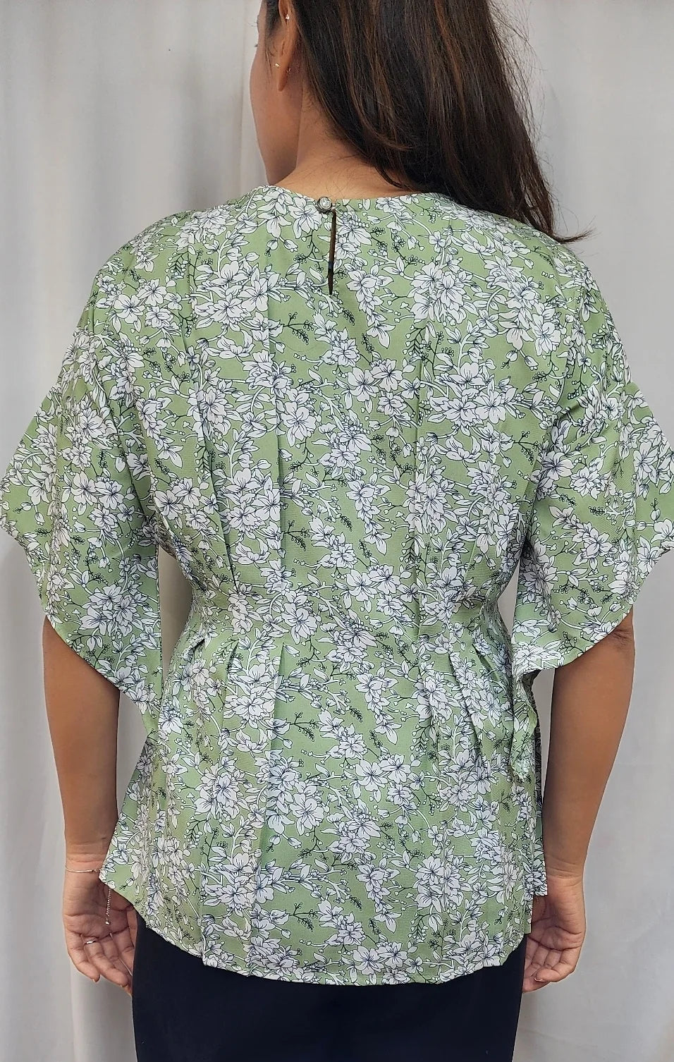 A woman viewed from the back wearing a light green short-sleeve blouse with a white floral pattern and dark navy pants, standing against a light gray curtain background.