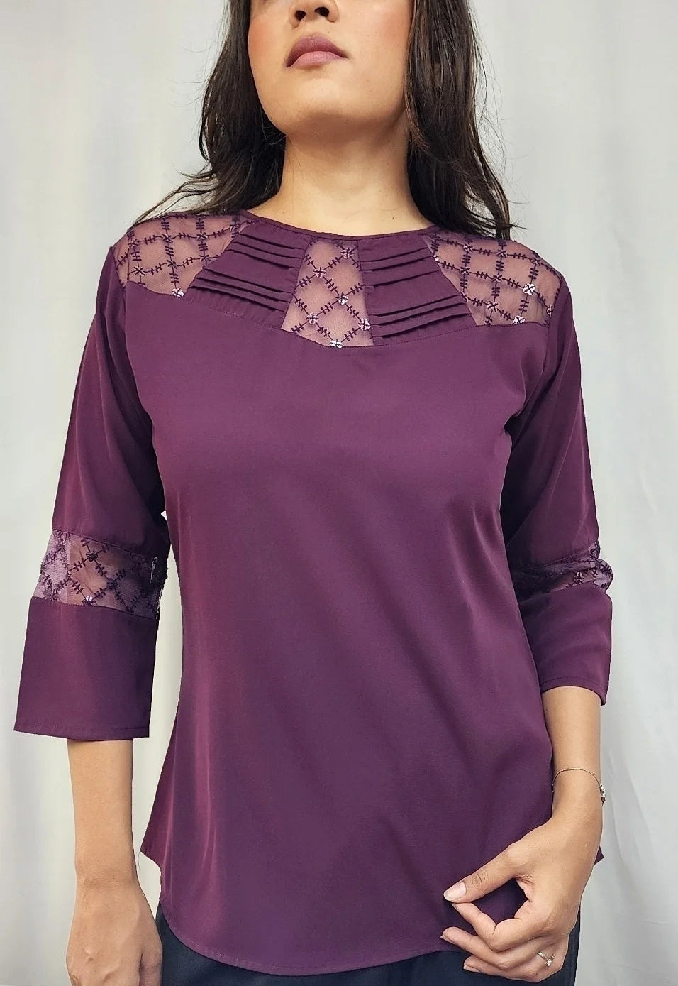 A woman wearing a purple three-quarter sleeve top with lace detailing on the shoulders and cuffs, paired with dark pants, standing against a light gray background.
