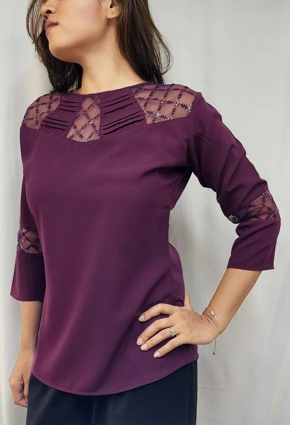 A woman wearing a purple three-quarter sleeve top with lace detailing on the shoulders and cuffs, paired with dark pants, standing against a light gray background.