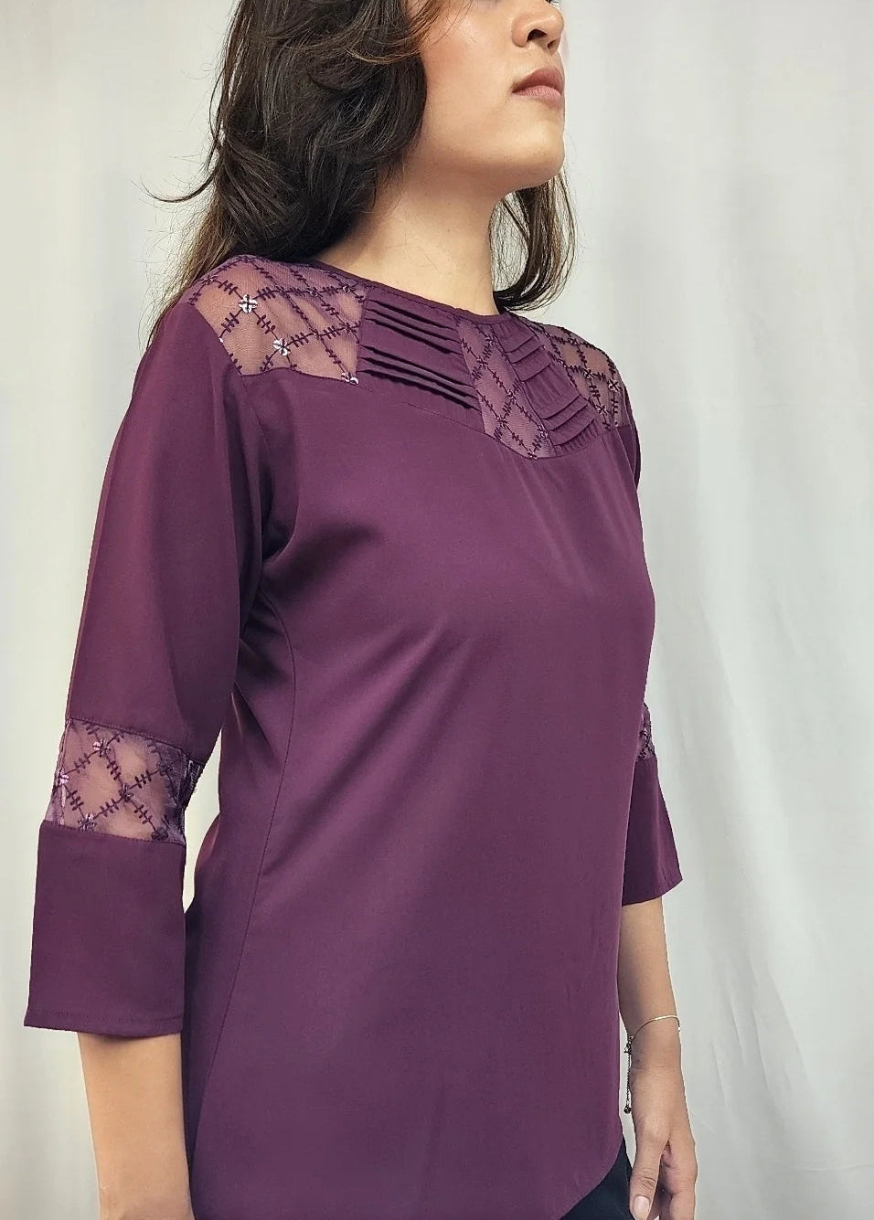 A woman wearing a purple three-quarter sleeve top with lace detailing on the shoulders and cuffs, photographed against a light neutral background.