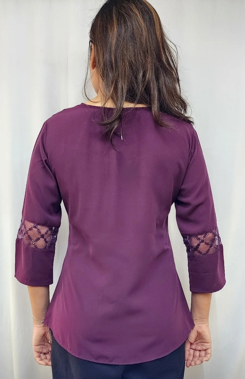 A woman viewed from the back wearing a purple three-quarter sleeve top with lace cutout details on the sleeves, paired with dark pants, set against a plain light background.