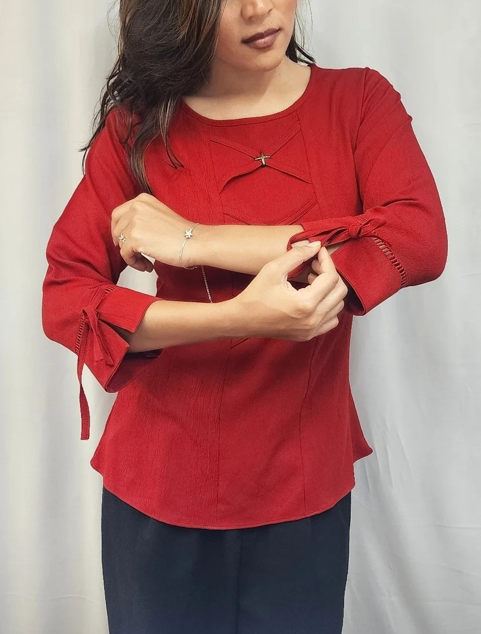 A woman wearing a red long-sleeve blouse with three-quarter sleeves and dark navy pants, adjusting the sleeve of the blouse against a light gray fabric backdrop.