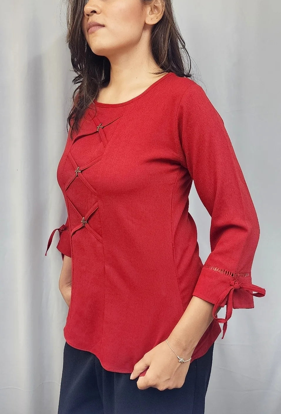 A woman modeling a red long-sleeve top with asymmetrical tie closures and side ties, paired with dark navy pants, standing against a light gray curtain backdrop.