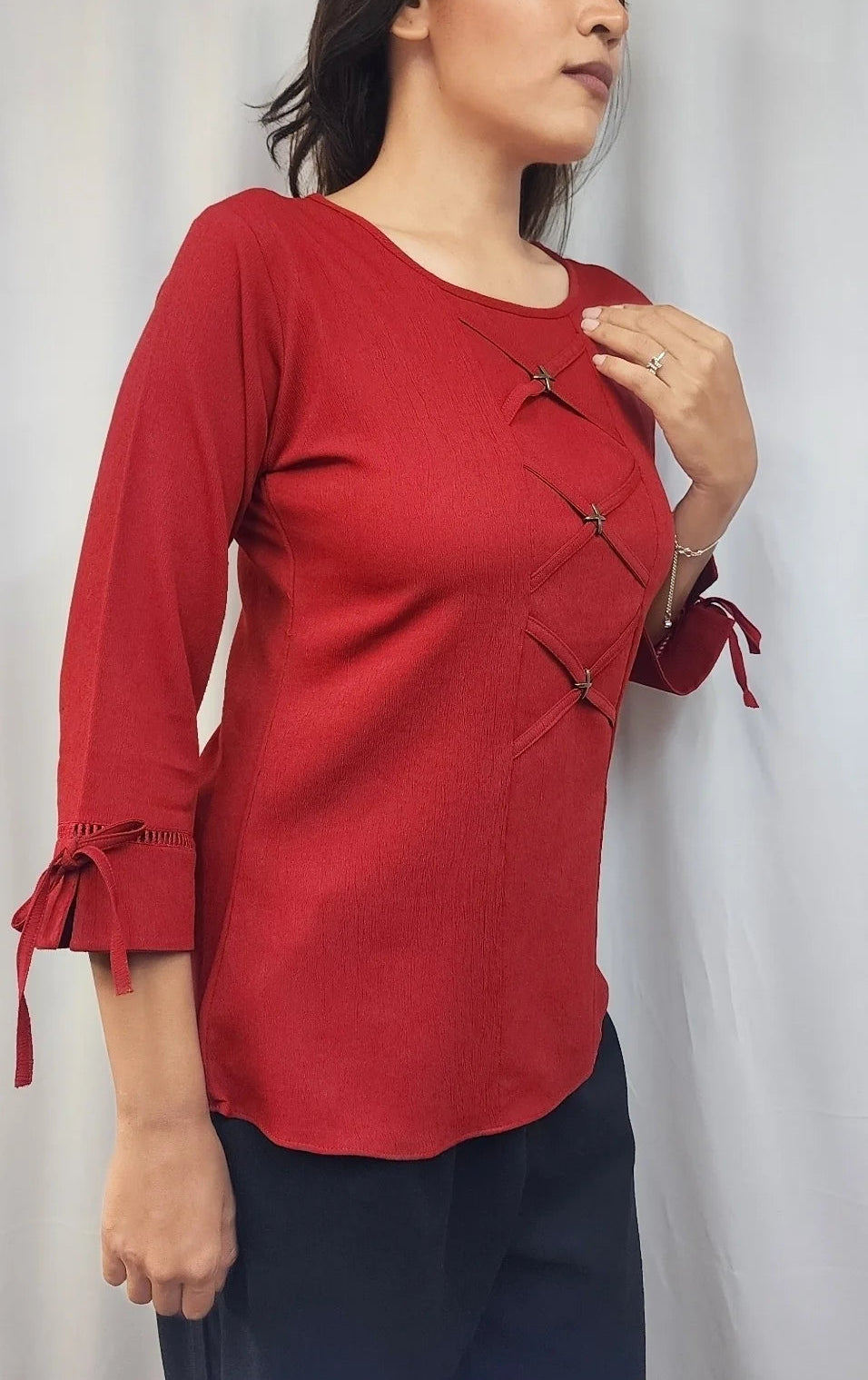 A woman wearing a red three-quarter sleeve top with dark buttons and side ties, paired with dark pants, standing against a light gray curtain backdrop.