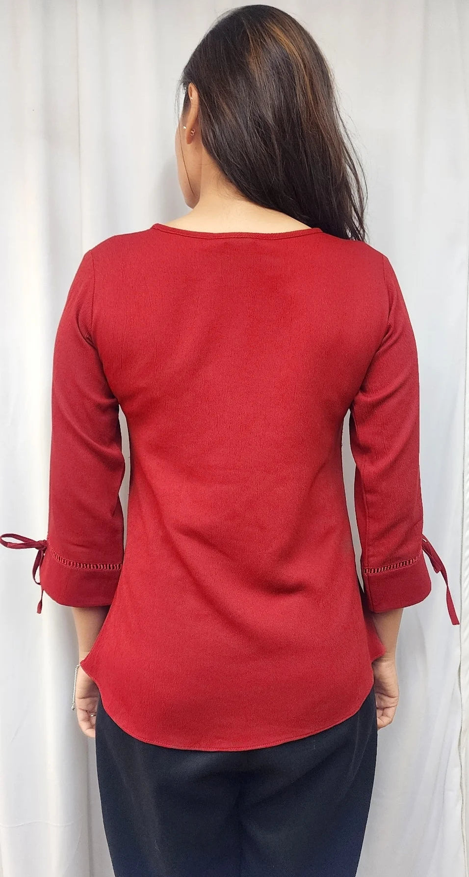 A woman viewed from behind wearing a red long-sleeve top with tie details on the cuffs and dark blue pants, standing in front of a white curtain.