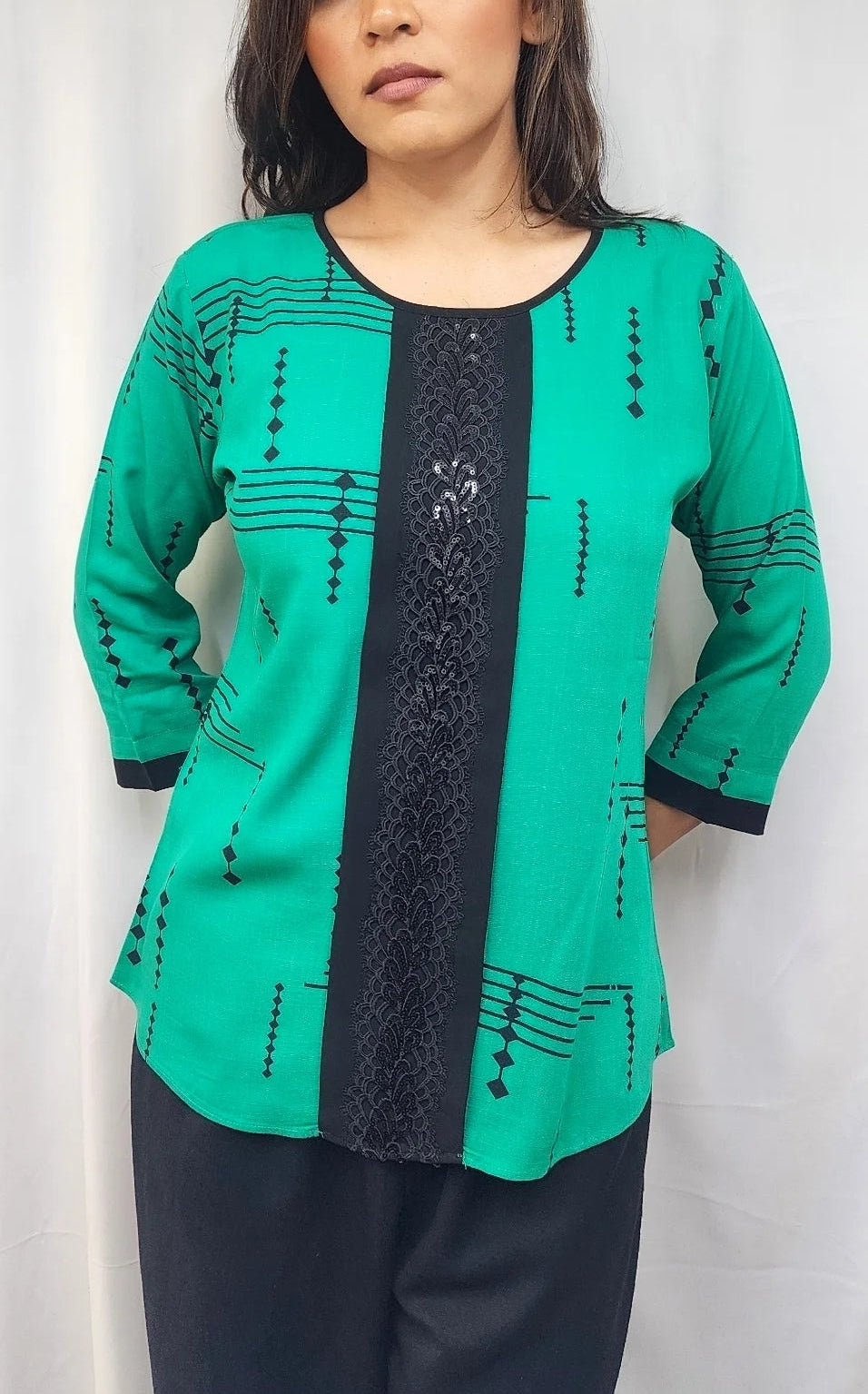 A woman wearing a green long-sleeve tunic top with a black vertical lace panel and black geometric patterns, paired with dark pants, standing against a white curtain background.