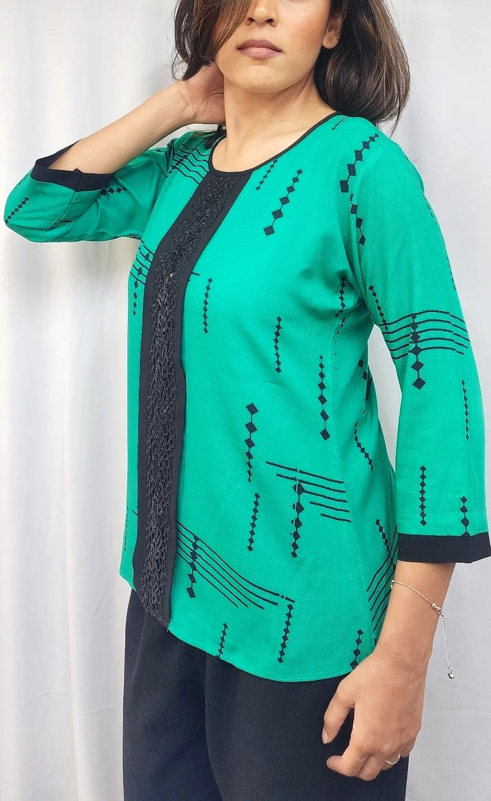 A woman modeling a bright green three-quarter sleeve top with black printed patterns and black lace trim on the front and cuffs, paired with black pants, set against a white curtain background.
