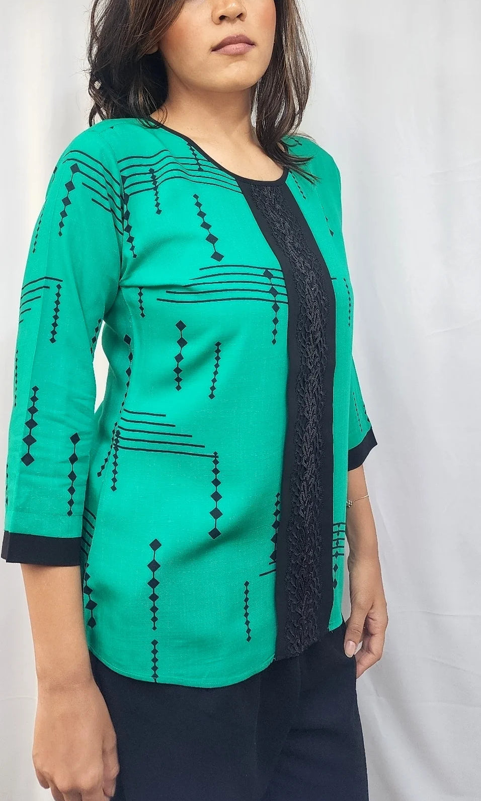 A woman modeling a teal green three-quarter sleeve top with black geometric patterns and a black lace-trimmed V-neckline, paired with dark pants, set against a white curtain background.