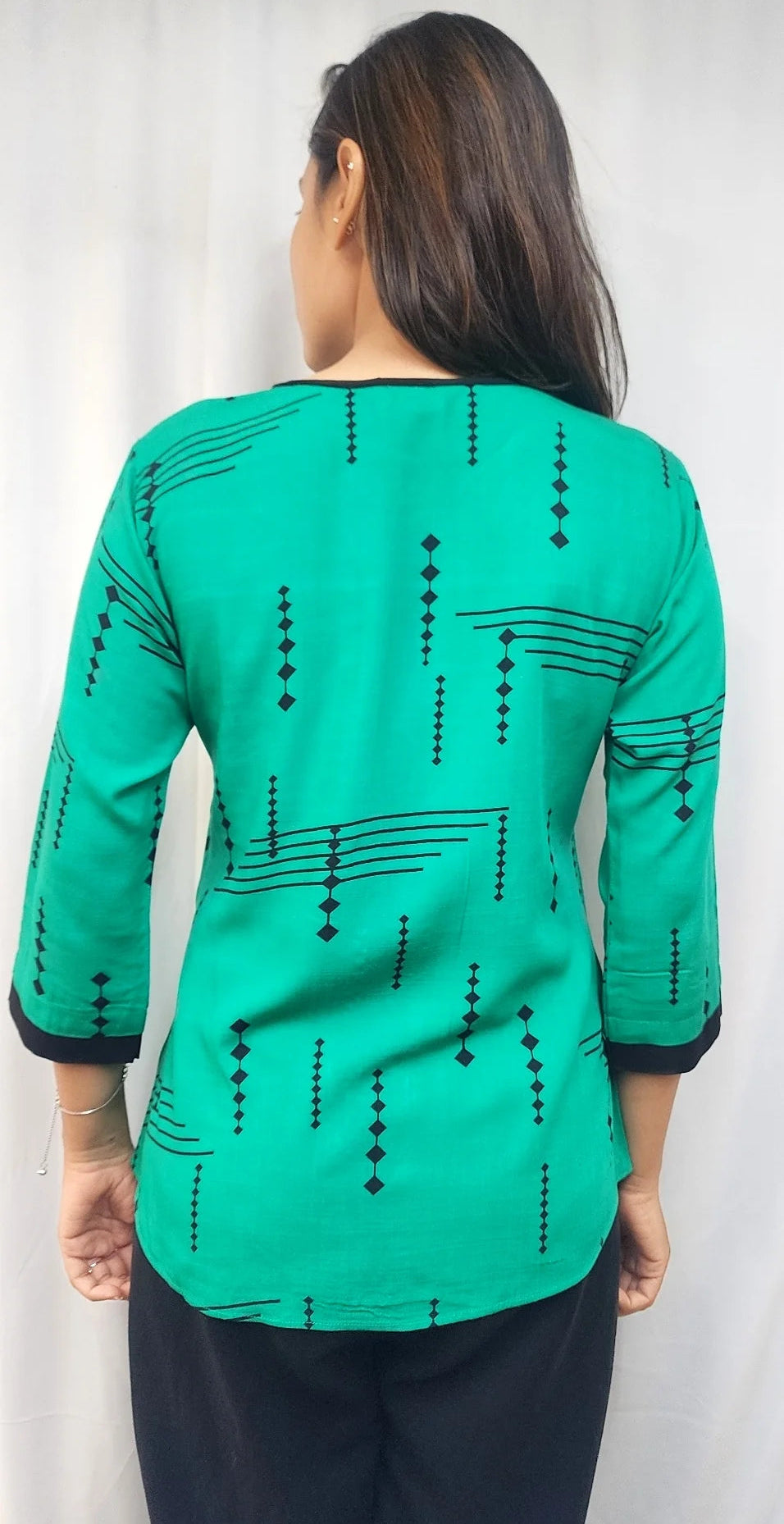 A woman viewed from the back wearing a bright green three-quarter sleeve top with black geometric patterns, paired with black pants, against a light gray curtain background.