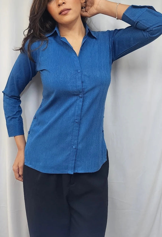 A woman modeling a blue long-sleeve button-up shirt with dark navy pants, standing against a light gray curtain backdrop.