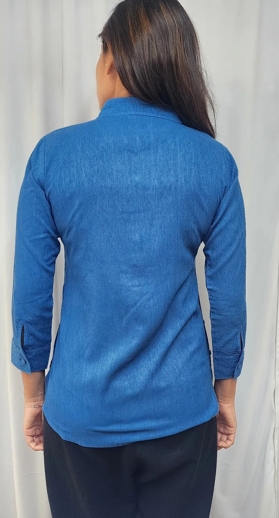A woman viewed from the back wearing a textured blue long-sleeve shirt and dark pants, standing in front of a light-colored curtain.