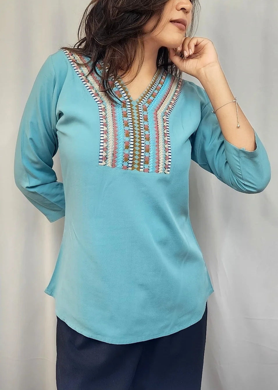 A woman wearing a light blue three-quarter sleeve kurta with colorful embroidered detailing around the V-neck, paired with dark navy pants, standing against a light gray curtain backdrop.