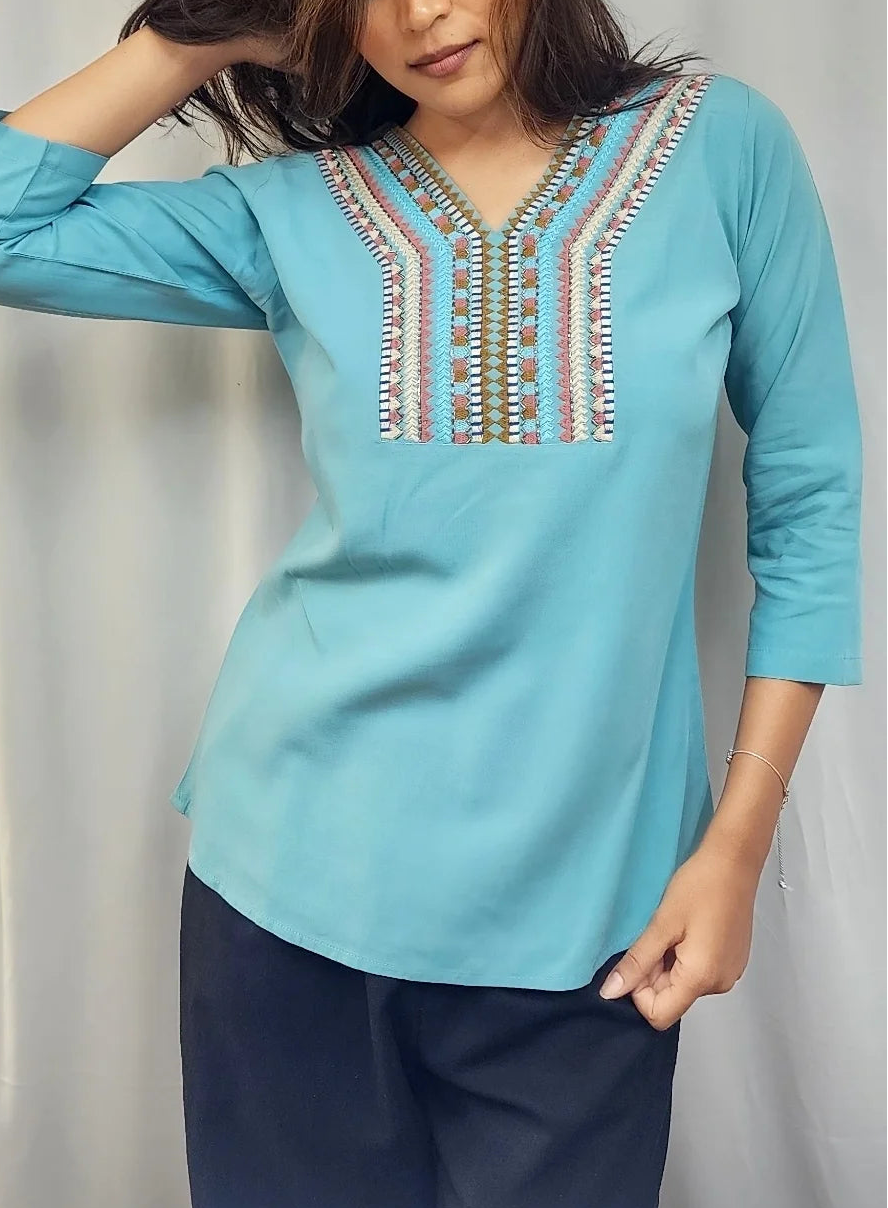A woman wearing a light blue three-quarter sleeve kurta with intricate multicolor embroidery around the V-neck, paired with dark navy pants, standing in front of a white curtain.