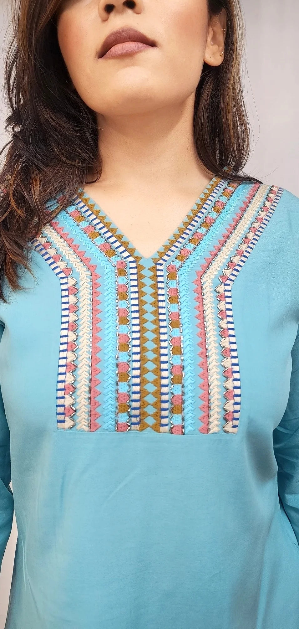 A close-up of a woman wearing a light blue long-sleeve tunic top with a colorful geometric embroidered neckline.