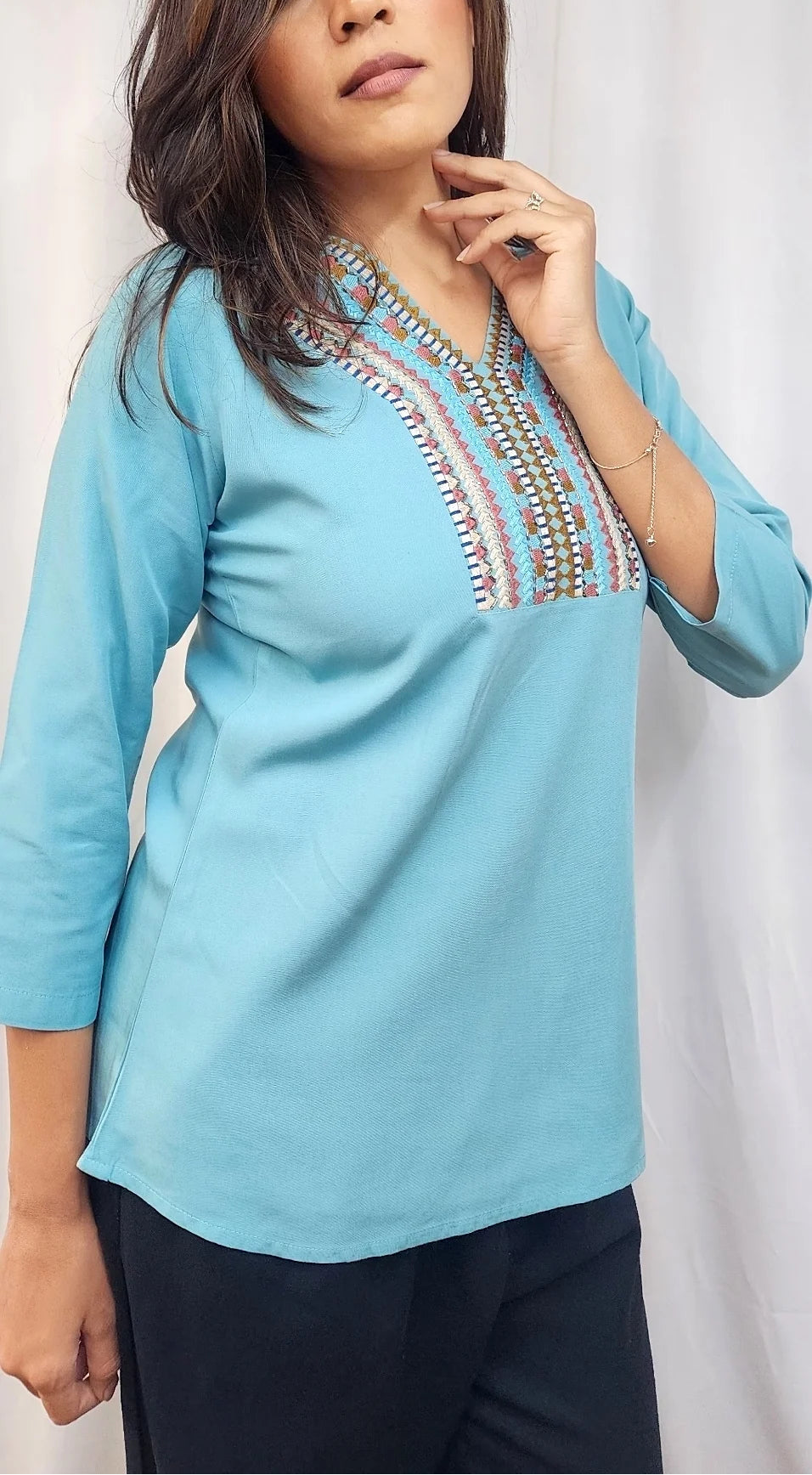 A woman wearing a light blue three-quarter sleeve kurta with colorful embroidered detailing around the V-neckline, paired with dark pants, posed against a white curtain backdrop.