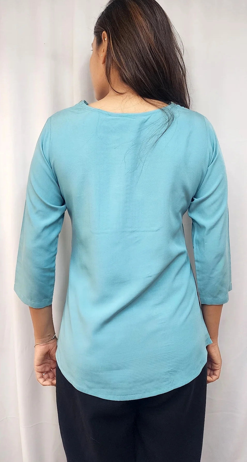 A woman viewed from the back wearing a light blue three-quarter sleeve top and black pants, standing in front of a white curtain.