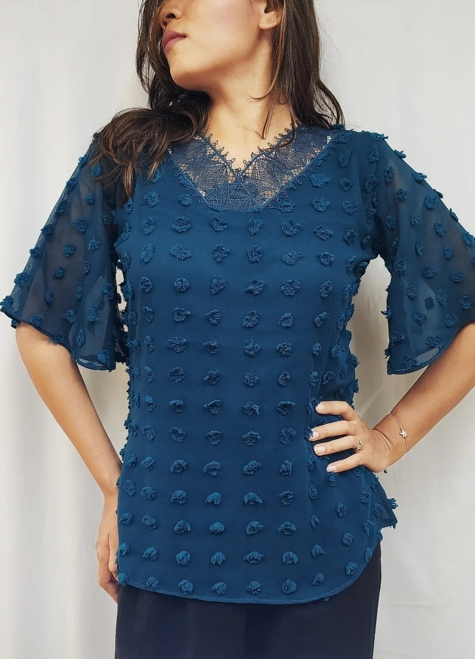 A woman wearing a textured blue short-sleeve blouse with a lace V-neckline, standing against a light gray background.
