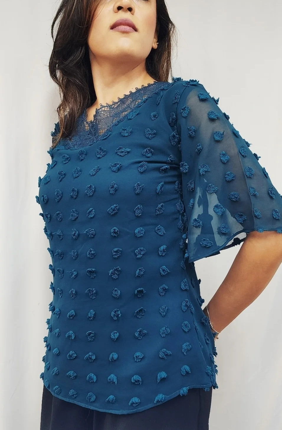 A woman wearing a teal blue textured sheer blouse with short flutter sleeves and a lace V-neckline, paired with dark navy trousers, set against a plain white background.